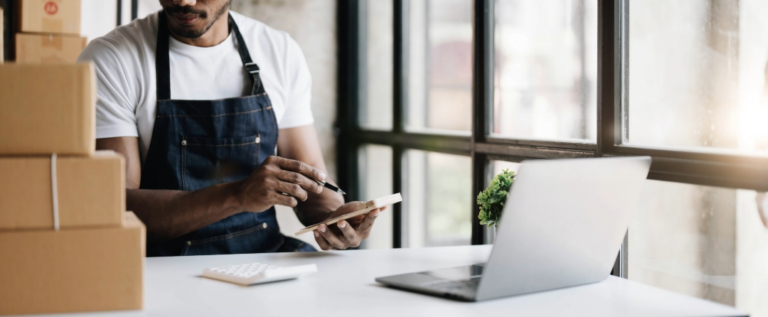 Small business owner using laptop and notepad to manage orders near stacked shipping boxes